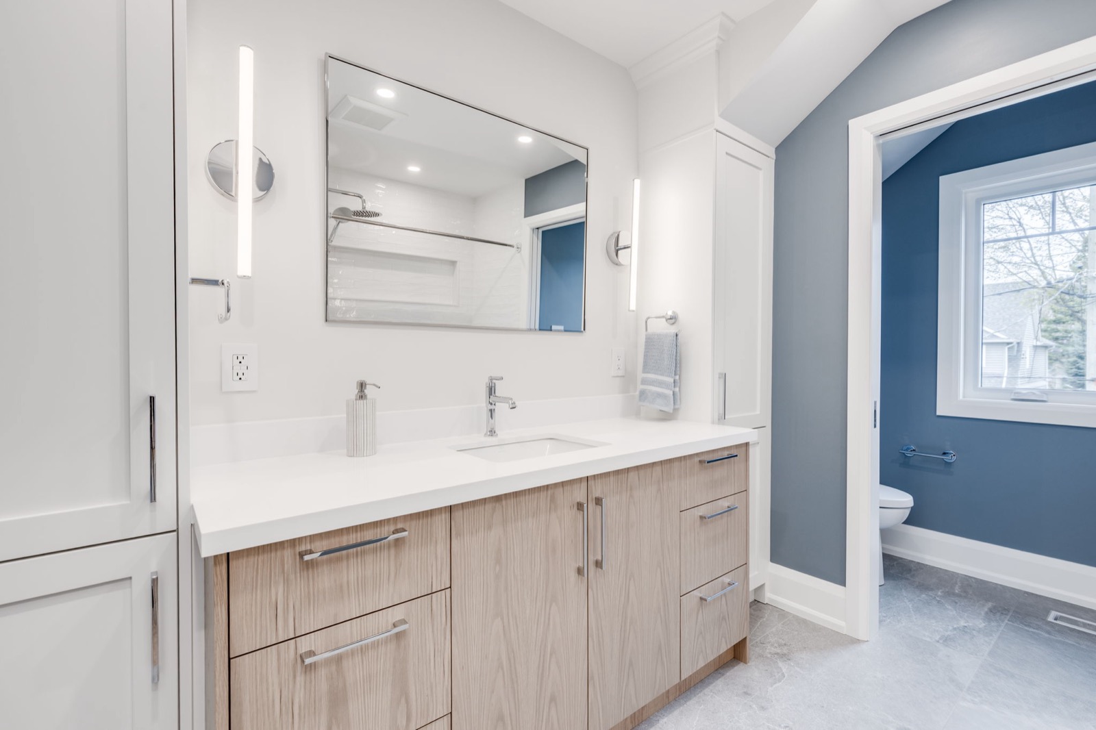Finished bathroom with a light wood vanity, white counters, chrome sconces, and blue grey wall colour.