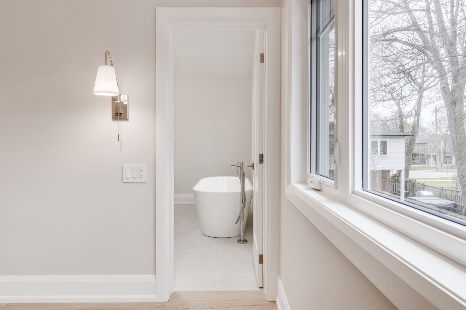 Bright warm white bathroom with freestanding tub, chrome filler, large window, and natural oak flooring.