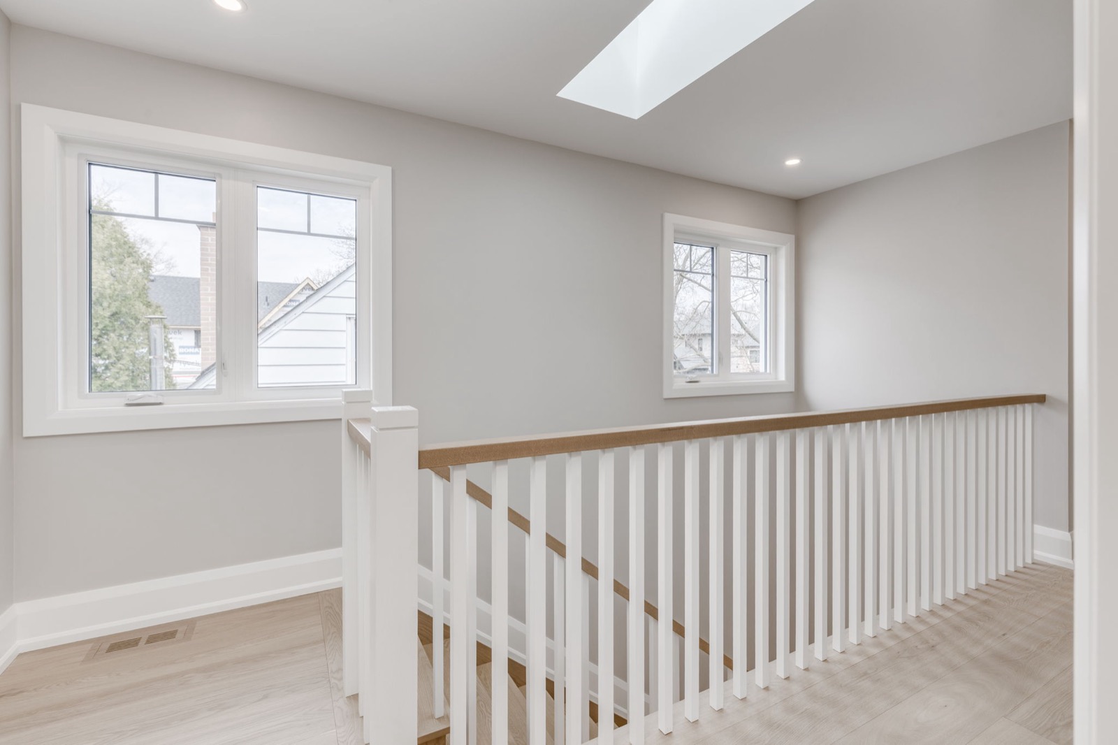 Bright upper hallway with white stair railing, oak floors, grey walls, windows and a skylight.